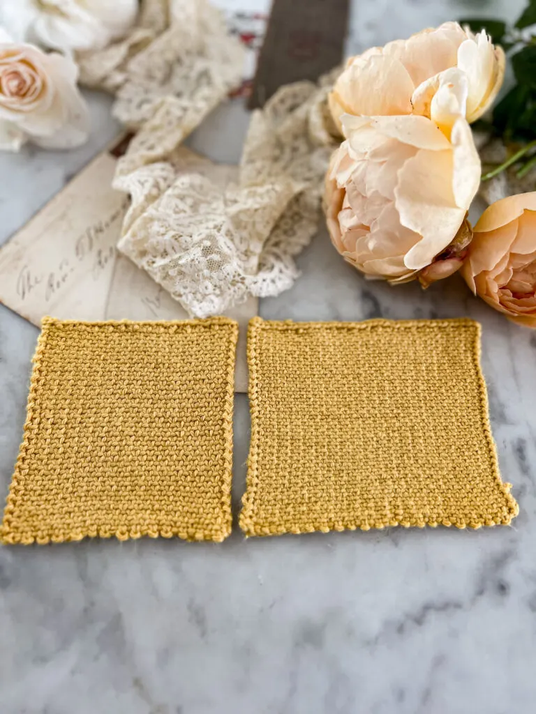 Two yellow swatches knit in linen stitch are laid out side by side on a white marble countertop. The left swatch was knit on larger needles and so the stitch pattern is visibly looser. Blurred in the background are white and peach roses, antique lace, and paper ephemera.