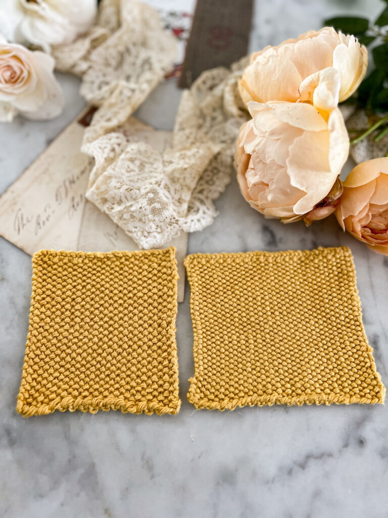 Two yellow swatches are laid out side by side on a white marble countertop. They're knit in linen stitch, but they're flipped over to show the back. The left swatch was knit with larger needles and therefore has larger, looser stitches. They're surrounded by white and peach roses, antique lace, and paper ephemera.
