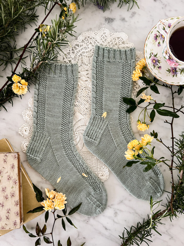The Madeleine Socks, a pair of mint-green handknit socks, are laid flat on a white marble countertop with both toes pointing to the right. The socks feature seed stitch and dainty cables running down the front and back. They're surrounded by vintage books, a teacup full of black tea, and yellow miniature roses.