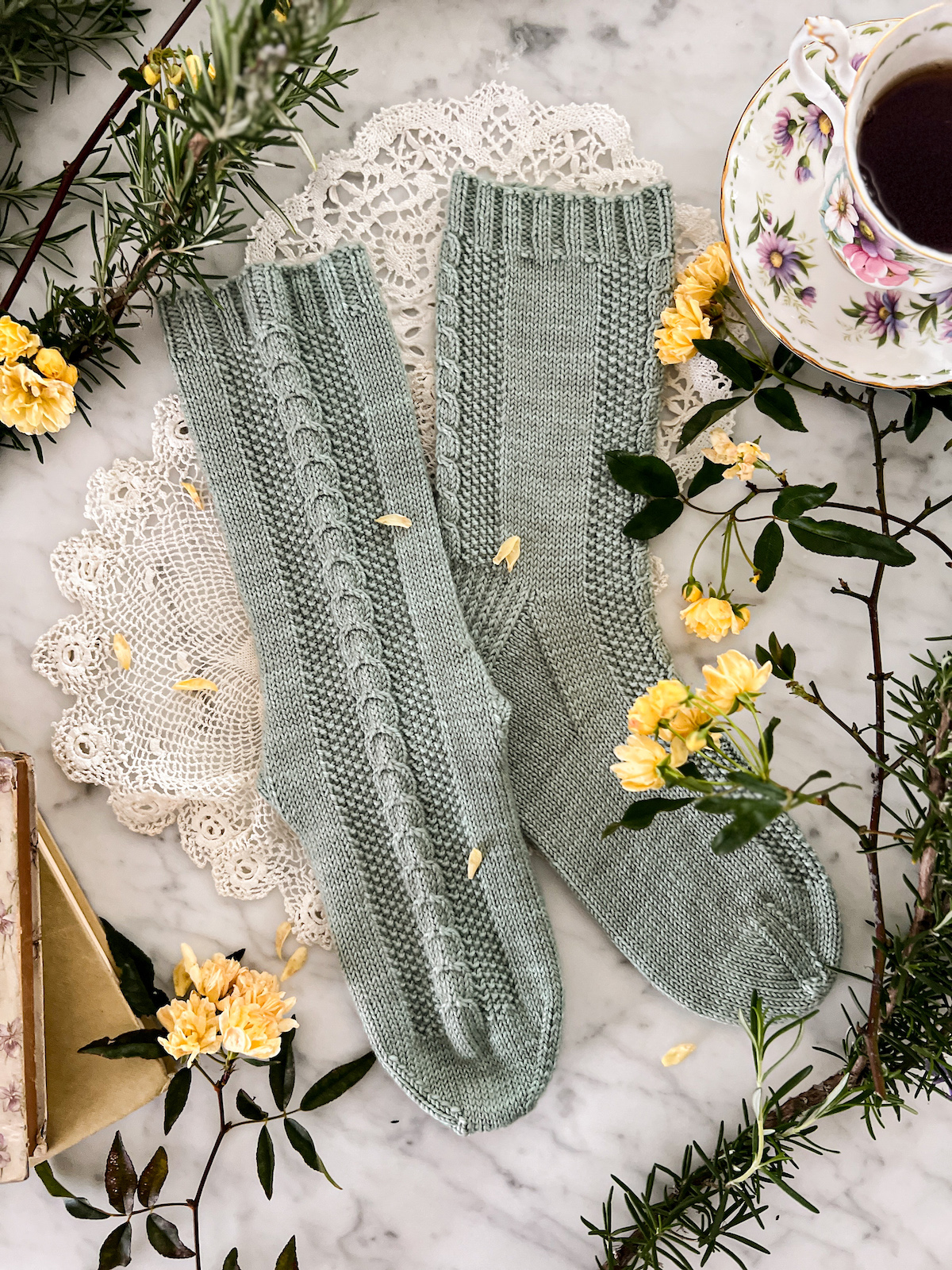A flatlay photograph of the Madeleine Socks with one sock on its side and one sock with the front panel displayed so the dainty cables are more visible. They're surrounded by antique books, a teacup full of black tea, and some miniature yellow roses.