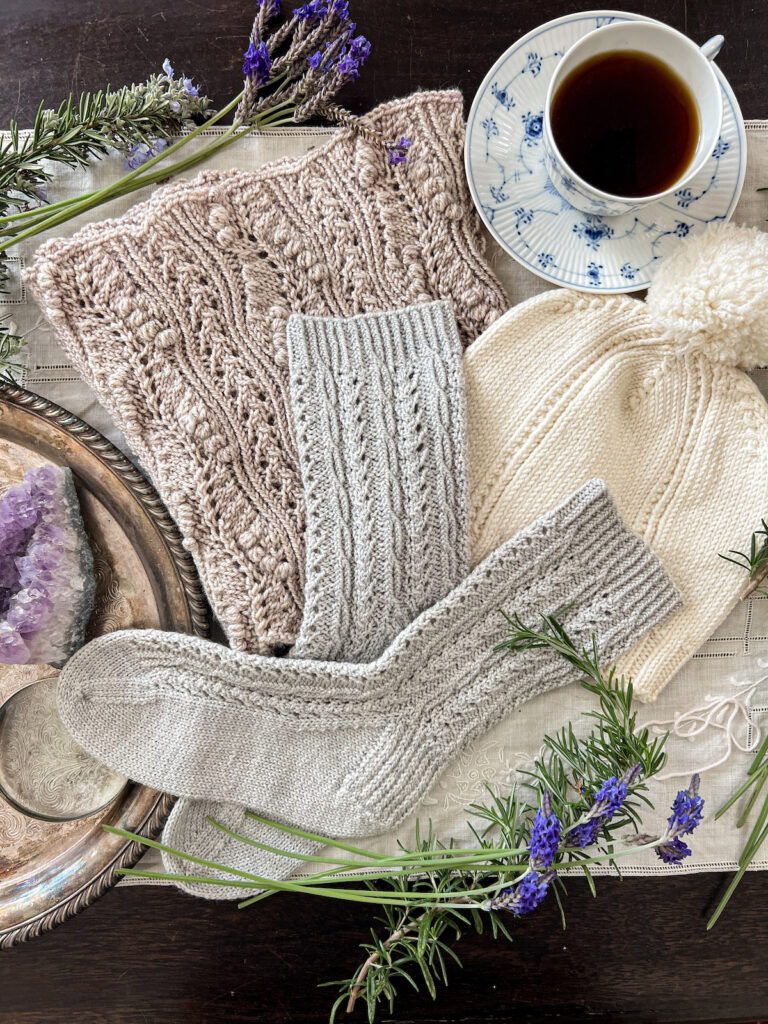 A hat, pair of socks, and cowl, all featuring twisted stitches as part of their stitch patterns, are laid on top of each other on an embroidered linen table cloth. They're surrounded by fresh greenery from the garden, a blue and white teacup full of black coffee, and a silver tray with some curios on it