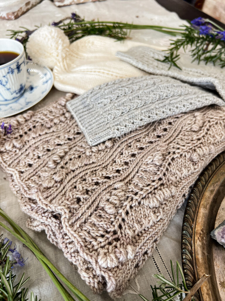 A close-up of the texture of a handknit cowl, showing the many twisted stitches in its stitch pattern. There are also eyelets and tiny crocheted bobbles.