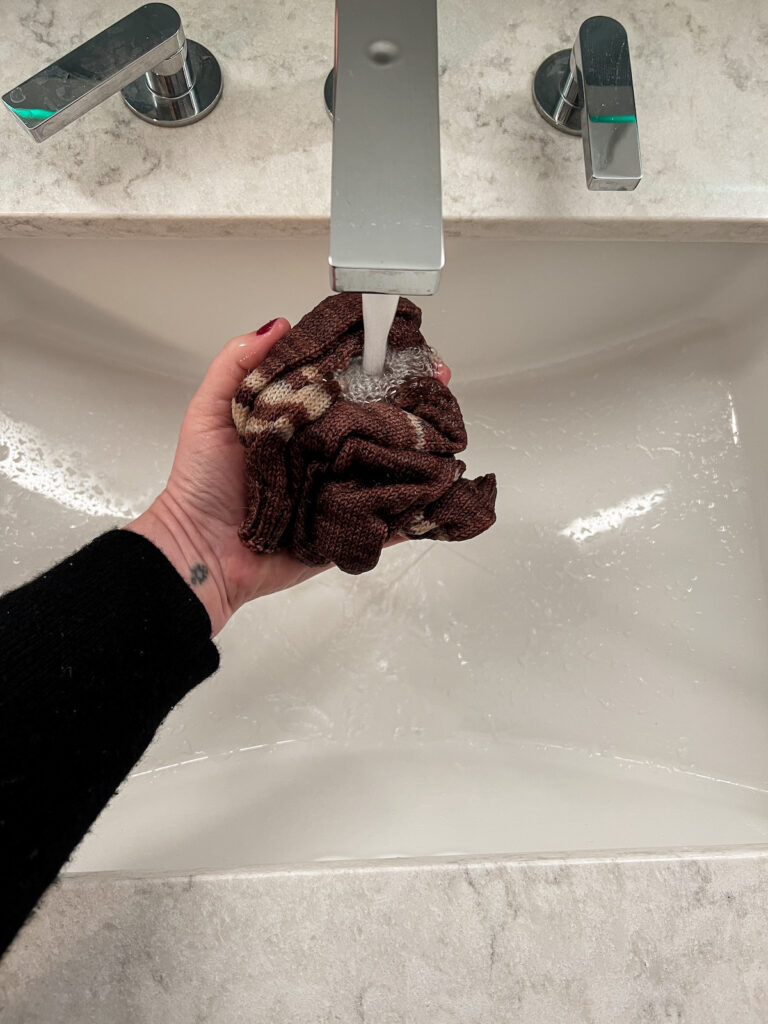 A white woman's left hand (mine!) cradles a pair of handknit socks in a hotel bathroom sink while fresh water flows over them.