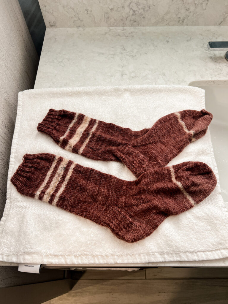 A freshly washed pair of pink, hand-knit socks with cream stripes sits on top of a dry towel.