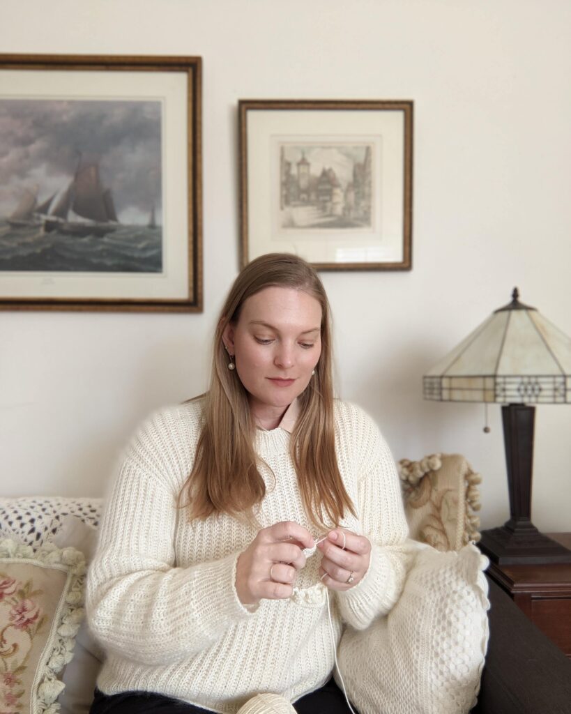 A white woman in her late 30s (me) wears a white fuzzy sweater while knitting on the sofa.