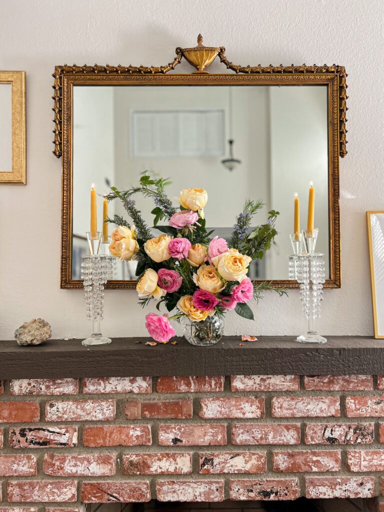 A crystal vase full of hot pink and yellow roses sits on a brown mantel above a brick fireplace. On either side is a crystal candlestick with a beeswax candle in it. An ornate gilt mirror is in the background.