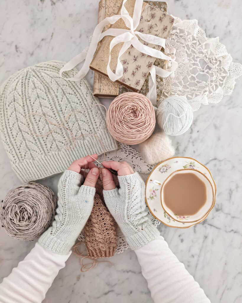 A white woman's hands (mine) are wearing pale blue fingerless mitts and knitting a sock. Around them are a teacup full of milky tea, some antique books, a handknit lacy hat, and some balls of leftover yarn.