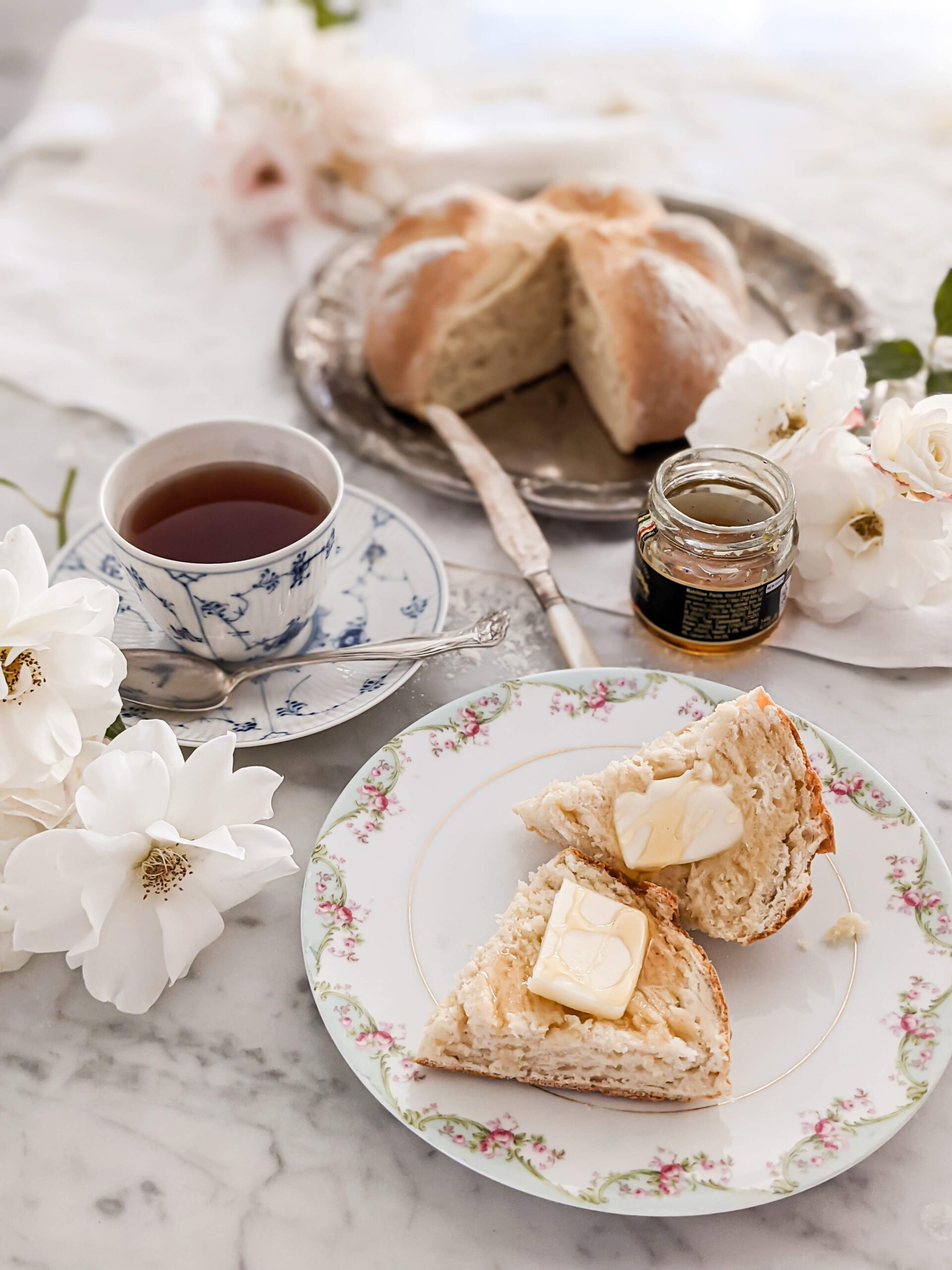 In the foreground is a floral plate with fresh bread and a teacup full of tea. Blurred in the background is the rest of the loaf of bread.