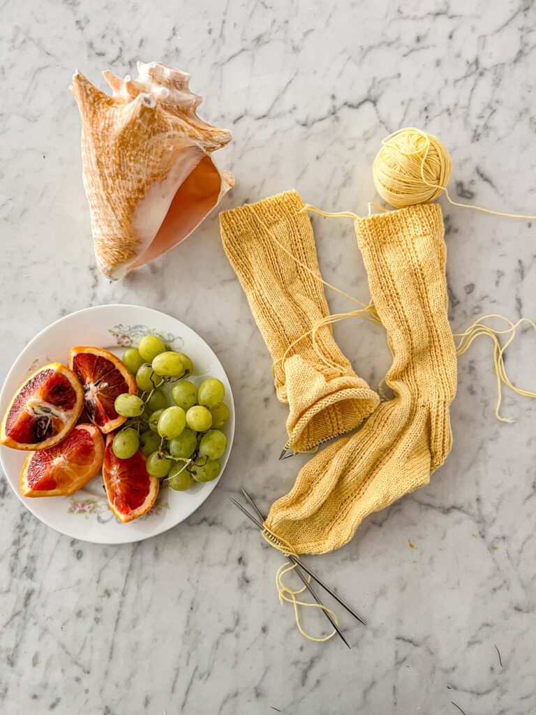 A pair of very yellow handknit socks in progress is crumpled on a white marble countertop next to a conch shell and a plate full of grapes and blood orange slices.