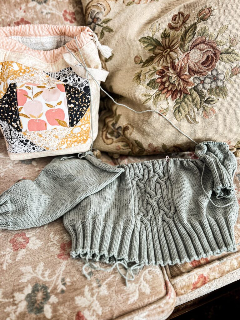 A pale green knit sweater in progress sits on a faded vintage sofa with floral upholstery. The sweater is a bottom-up raglan and the sleeves have just been attached. It has a cabled panel down the center. Above the sweater are a quilted project bag and a needlepoint pillow.