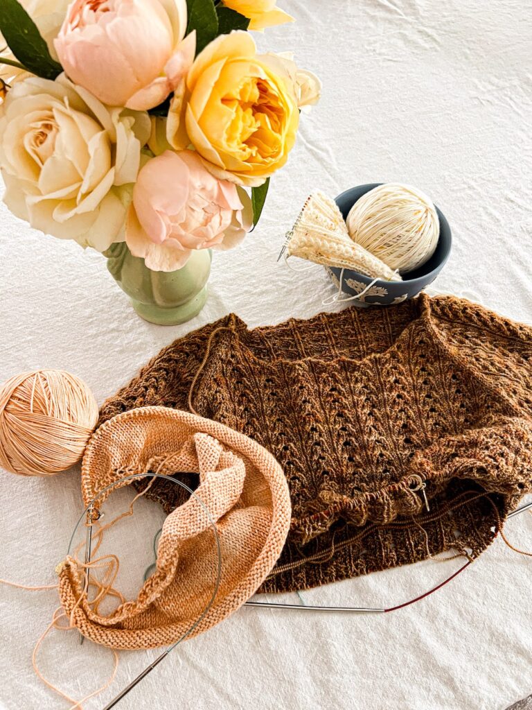 A top-down photo of a brown lace knit tshirt in progress. It has been knit down to just below where the sleeves were divided from the body. The t-shirt is sitting with a peach shawl in progress and a white sock with yellow speckles that is on a set of tiny circular needles. A vase of pastel roses is blurred at the top of the image.