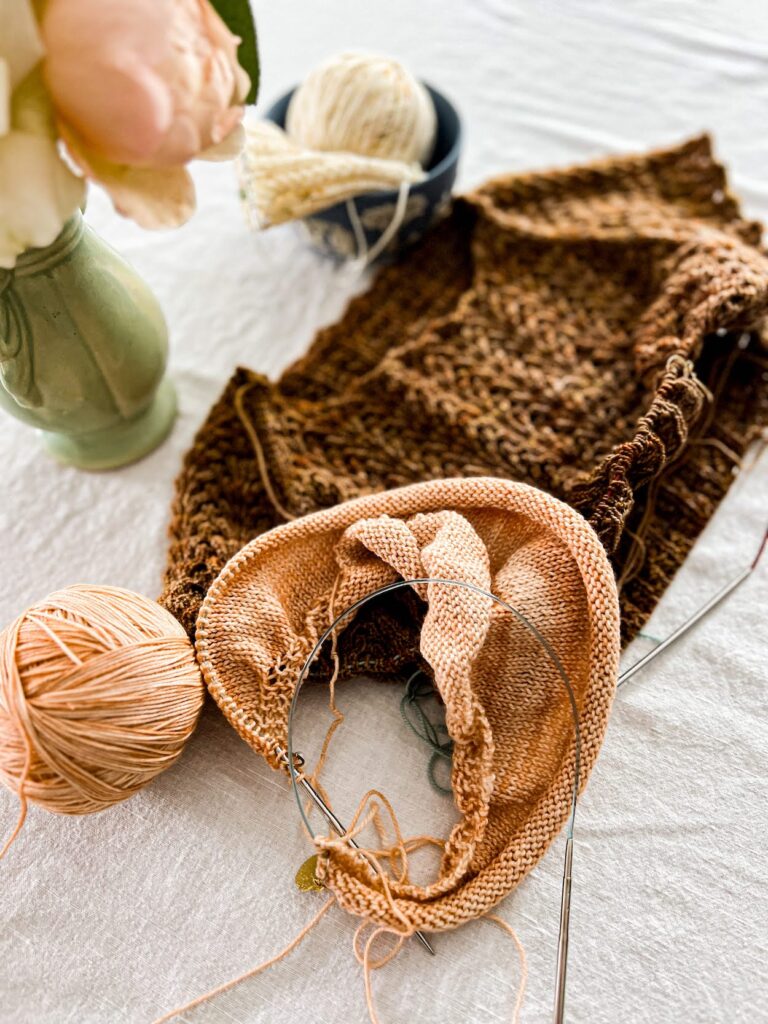 A photo of a peach stockinette shawl in progress on a set of long circular needles. Blurred in the background are other brown and cream knitting projects.