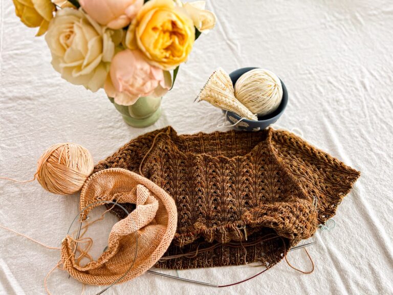 A top-down photo of a brown lace knit tshirt in progress. It has been knit down to just below where the sleeves were divided from the body. The t-shirt is sitting with a peach shawl in progress and a white sock with yellow speckles that is on a set of tiny circular needles. A vase of pastel roses is blurred at the top of the image.