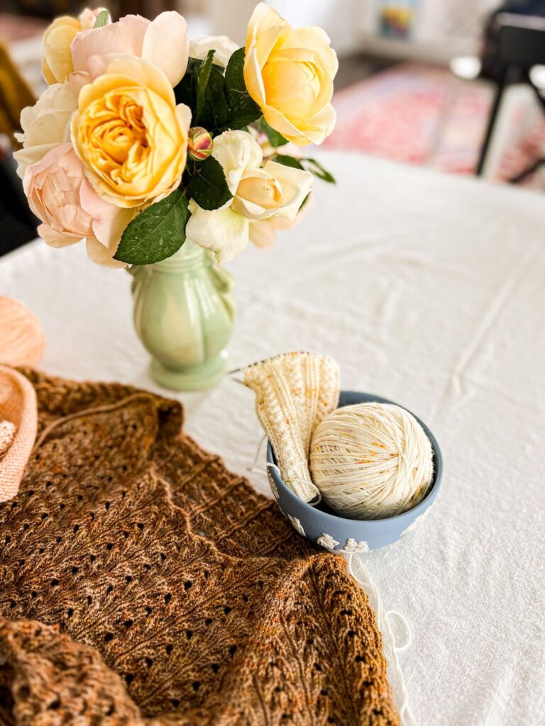 A photo of a brown lace knit tshirt in progress and a creamy sock in progress. The sock is in a light blue jasperware bowl along with the ball of yarn being used to knit it. A pale green vase full of pastel flowers fills the top left of the photo.