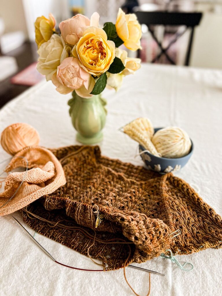 A photo of a brown lace tshirt in progress on a set of circular knitting needles. The photo is focused on the bottom hem where the shirt is growing. Blurred in the background are more knitting projects and a vase of pastel roses.