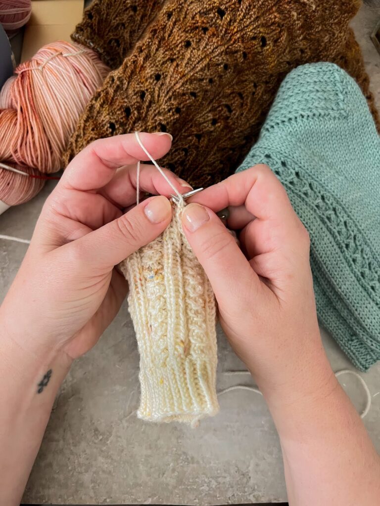 A white woman's hands working on a knit sock in progress showing how to tension the yarn in the left hand (continental style). In the background are several unfinished knitting projects.