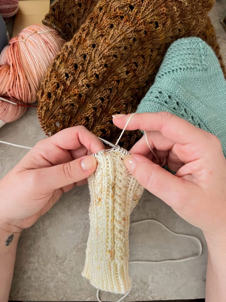 A white woman's hands working on a knit sock in progress showing how to tension the yarn in the right hand (English style). In the background are several unfinished knitting projects.