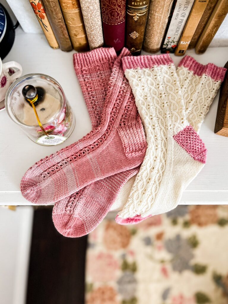 Two pairs of handknit socks sit on top of a white dresser surrounded by antique books and a candle under a glass cloche. The left pair of socks is pink with columns of cables running up the feet and legs. The right pair of socks is white with pink cuffs, heels, and toes, and is filled with lots of tiny twisted cables and columns of eyelets.