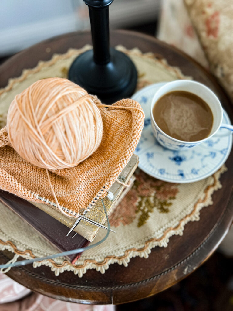 A pile of peach-colored knitting on a vintage side table with a blue and white teacup full of coffee next to it.