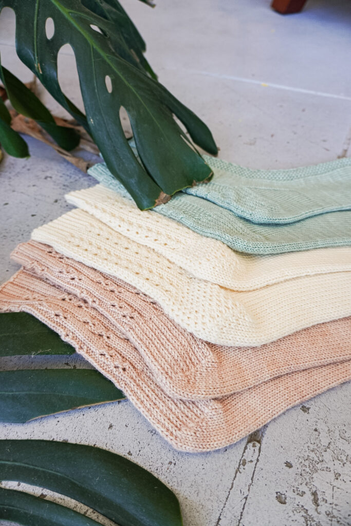 A low-angle photo of three pairs of socks laid flat on a gray concrete floor. In the foreground is a pink pair, with a cream pair in the middle and a light green pair furthest back. All three pairs of socks have textured heels that are highlighted in the middle of the photo.