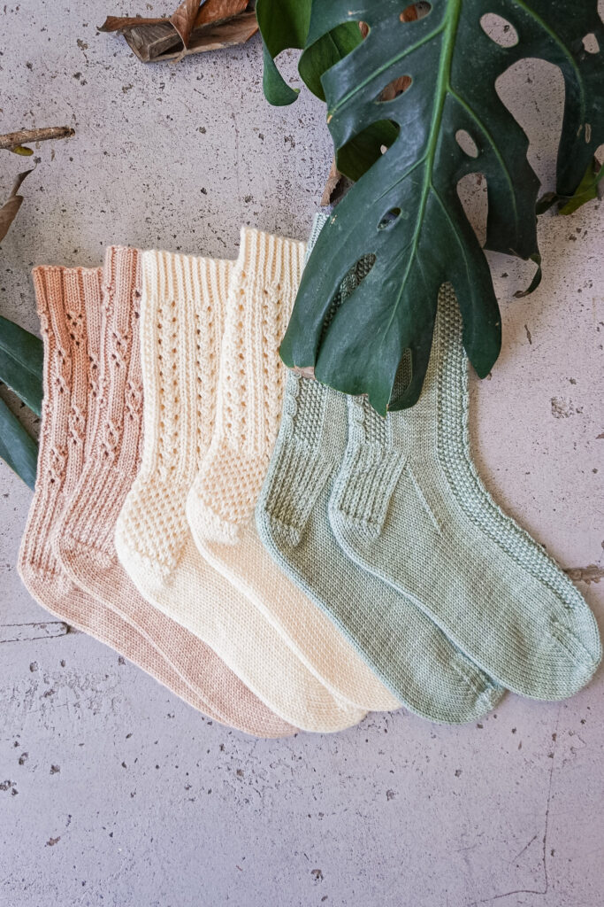 A top-down photo of three pairs of socks laid flat on a gray concrete floor. On the left is a pink pair, with a cream pair in the middle and a light green pair on the right. All three pairs of socks have textured heels.
