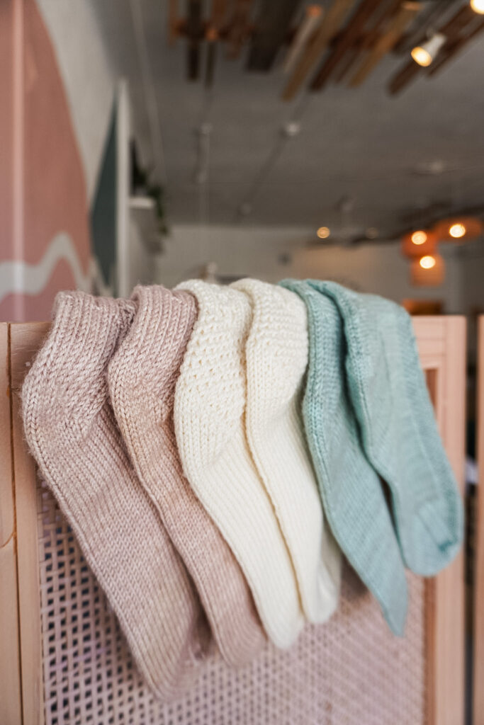 A side angle photo showing how three pairs of handknit socks are draped over the top of a wicker screen. The socks are draped so that their textured heels are visible. On the left is a pink pair, with a cream pair in the middle and a light green pair on the right.