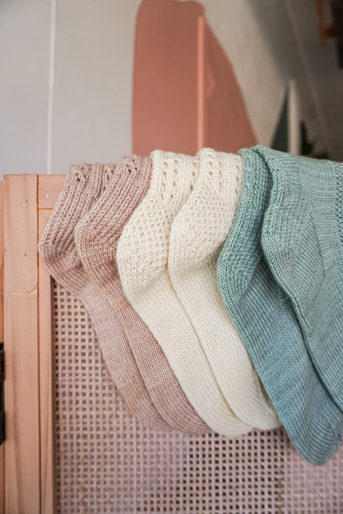 Three pairs of handknit socks are draped over the top of a wicker screen. The socks are draped so that their textured heels are visible. On the left is a pink pair, with a cream pair in the middle and a light green pair on the right.