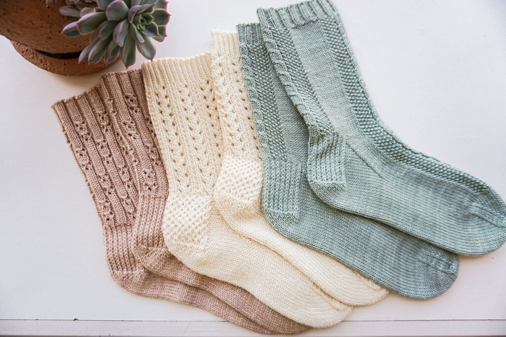 A horizontal photo taken from the top-down showing how three pairs of handknit socks with textured heels are laid out flat on a white countertop. On the left is a pink pair, with a cream pair in the middle, and a light green pair on the right.
