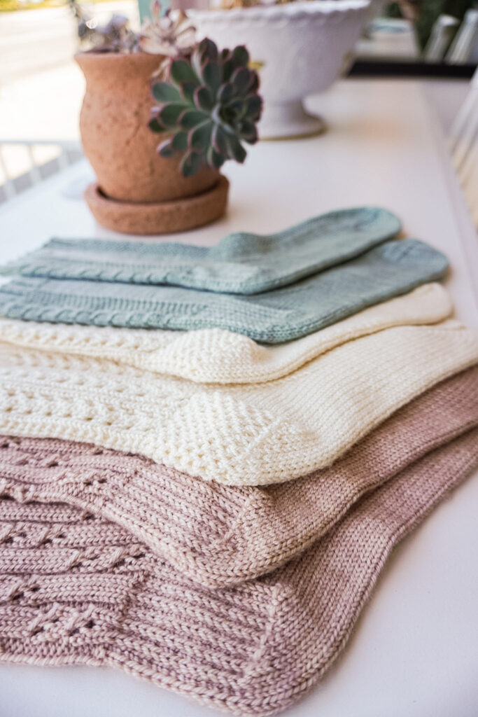 A vertical photo showing how three pairs of handknit socks with textured heels are laid out flat on a white countertop. In the foreground is a pink pair, with a cream pair in the middle, and a light green pair furthest back.
