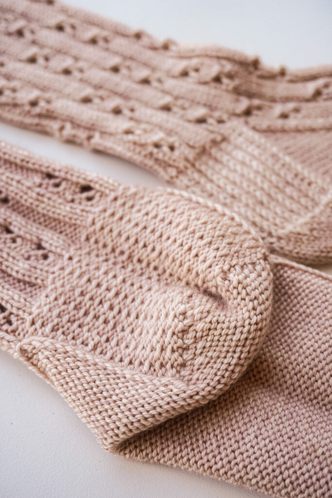 A close-up on the heels of a pink pair of handknit socks. In the foreground the sock is turned inside out to show the extra layer of cushioning provided by the slipped stitches in the heel. In the background, blurred slightly, the sock is left right-side out to show the columns of slipped stitches in the heel flap.