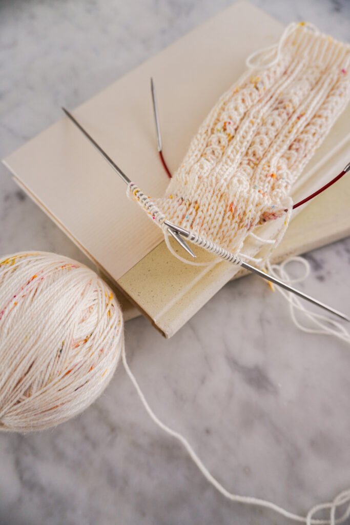 A cream sock in progress sits on top of a small stack of books on a white marble countertop. The sock is richly textured and has tiny orange, pink, and blue speckles.