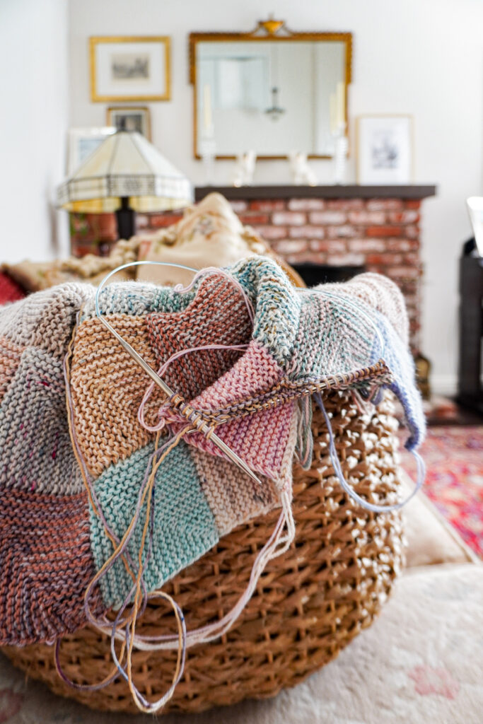 A woven basket with a multicolored mitered square blanket in progress poking over the top of the basket.