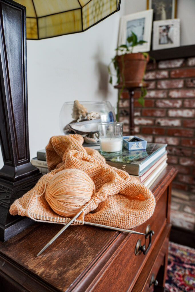 A side table with some coral-colored knitting on it. Blurred in the background are a stack of books and a glass bowl full of sea shells.