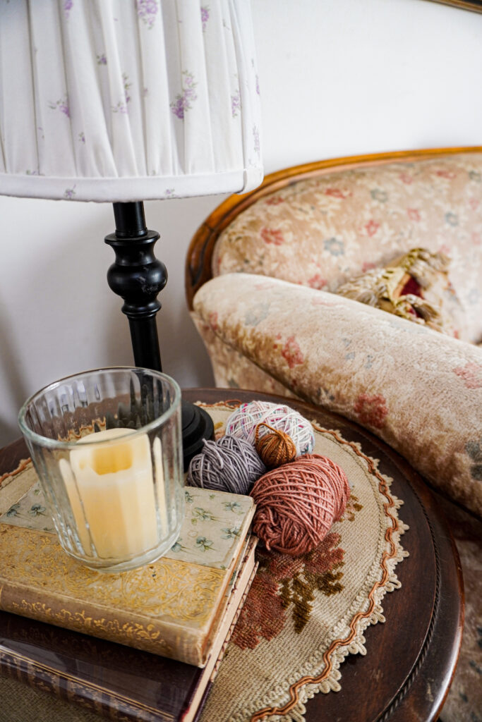 A side table with some antique books, a candle, a lamp, and several small balls of scrap yarn.