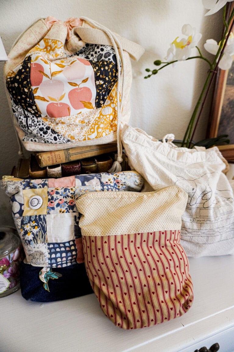A pile of several different project bags full of knitting stacked on a white dresser.