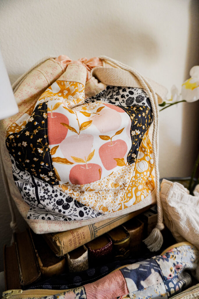 A close-up on a quilted project bag with a drawstring closure. The quilted part of the bag features fruit and autumnal foliage in shades of pink, goldenrod, tan, and black.
