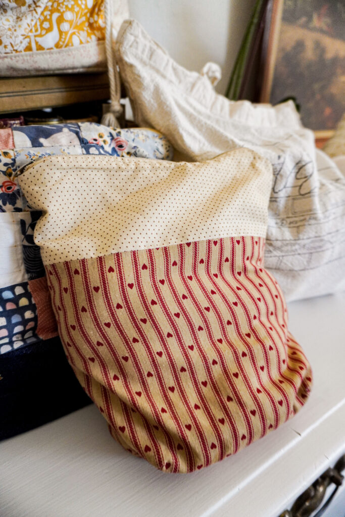 A close-up on a project bag made from two contrasting panels of fabric. The top panel is tan with tiny red polka dots. The bottom panel is a darker tan with red stripes and hearts.