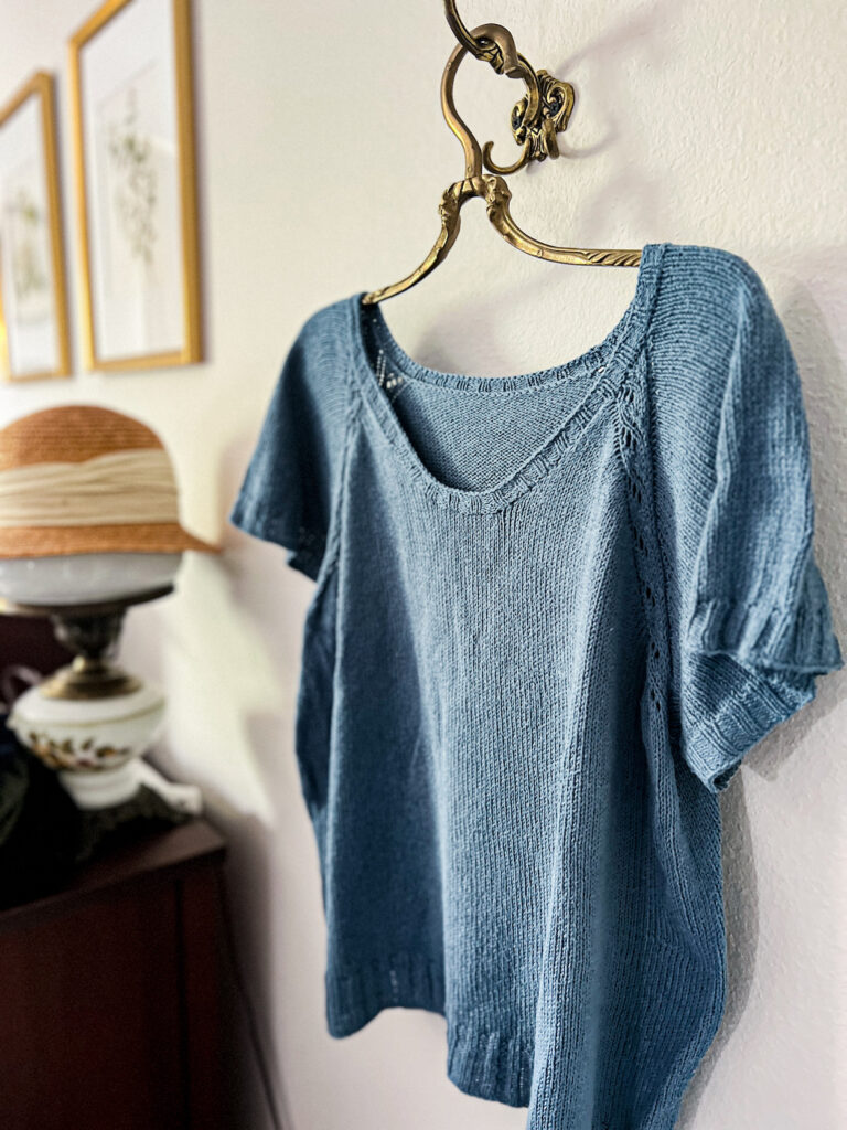 A light blue knit tshirt is hanging from a brass art nouveau clothes hanger. Do not store your silk or linen knits like this!