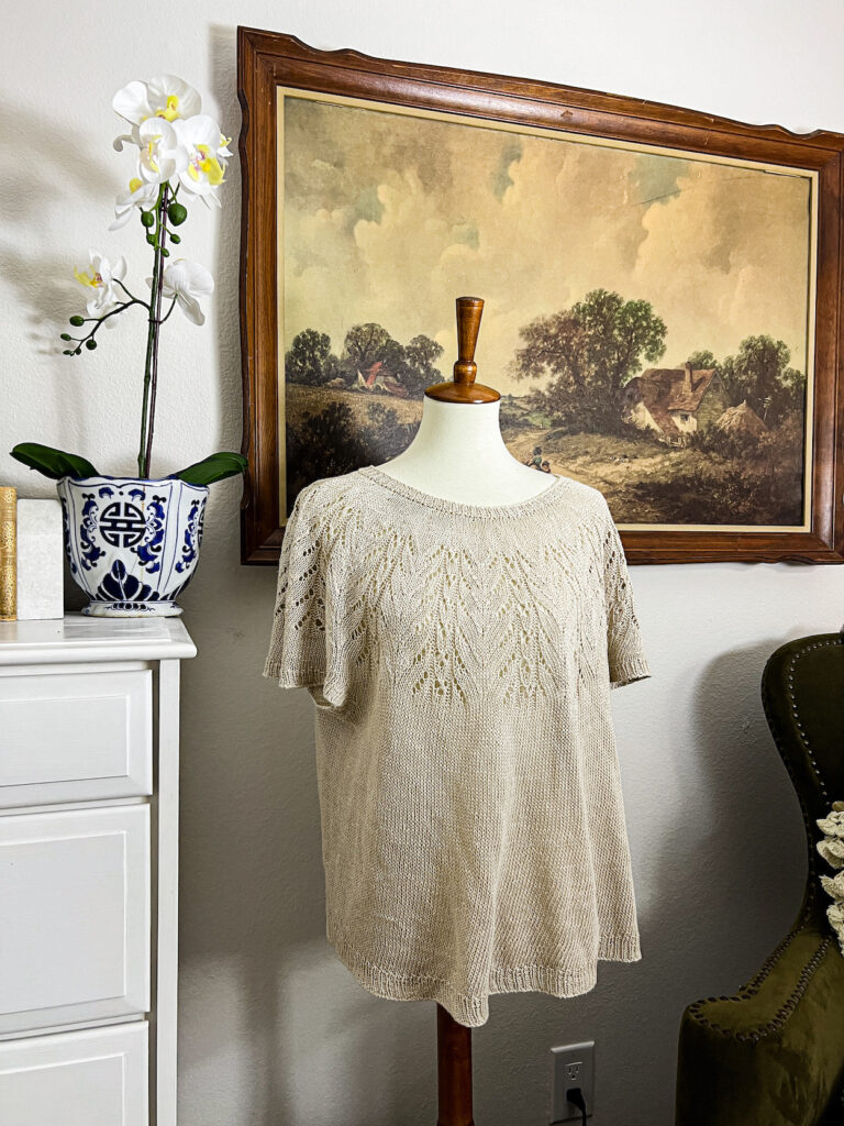 A tan knit tshirt with an intricate lace yoke is displayed on a white dressmaker's form next to a white dresser and in front of a vintage landscape print.