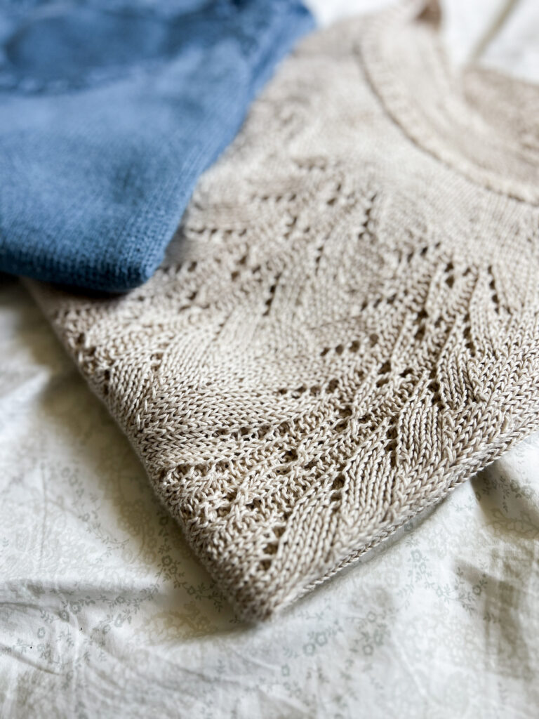 A folded tan knit top with intricate lace yoke fills the foreground of the photo. The top is knit with pure linen yarn.