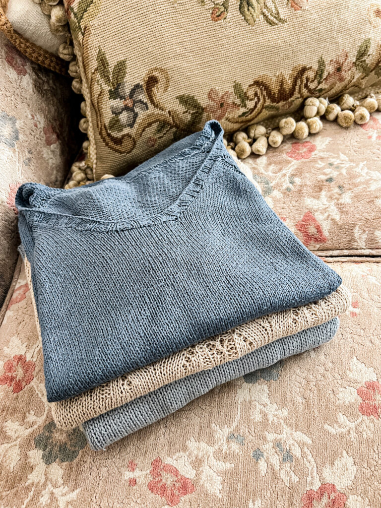 A folded stack of blue and tan knit tops sits on a faded floral sofa cushion.