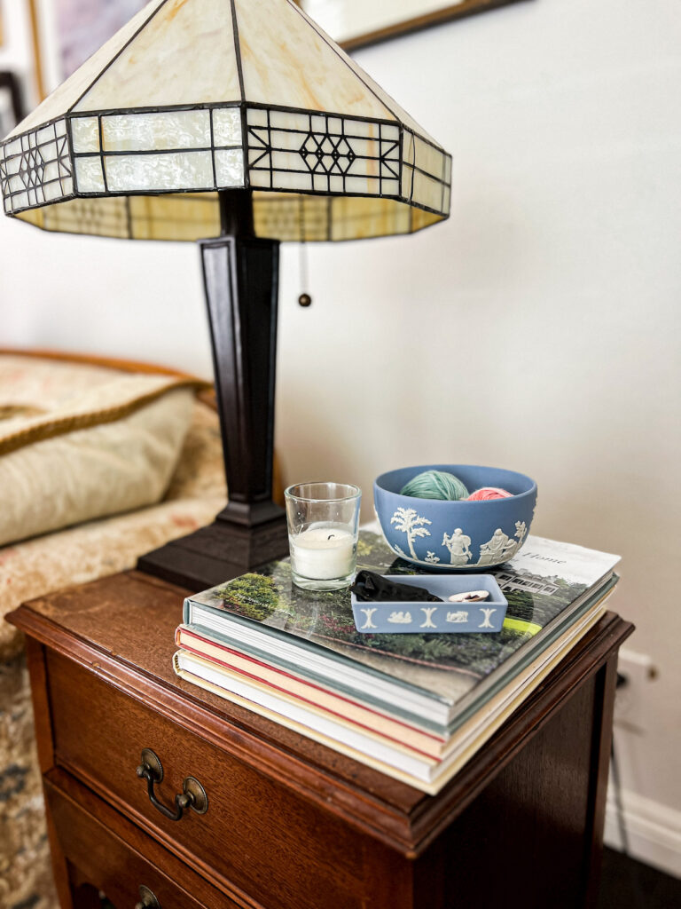 A light blue Jasperware bowl and lidded box sit on top of a pile of books next to a small tea candle. The bowl is full of small balls of yarn and the box holds tiny treasures from the beach. Next to them is a Tiffany-style lamp with a white shade.