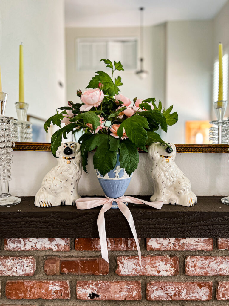 A straight-on photo of a light blue and white Jasperware vase full of pink roses and greenery sits on a dark brown mantel between two white Staffordshire dogs.