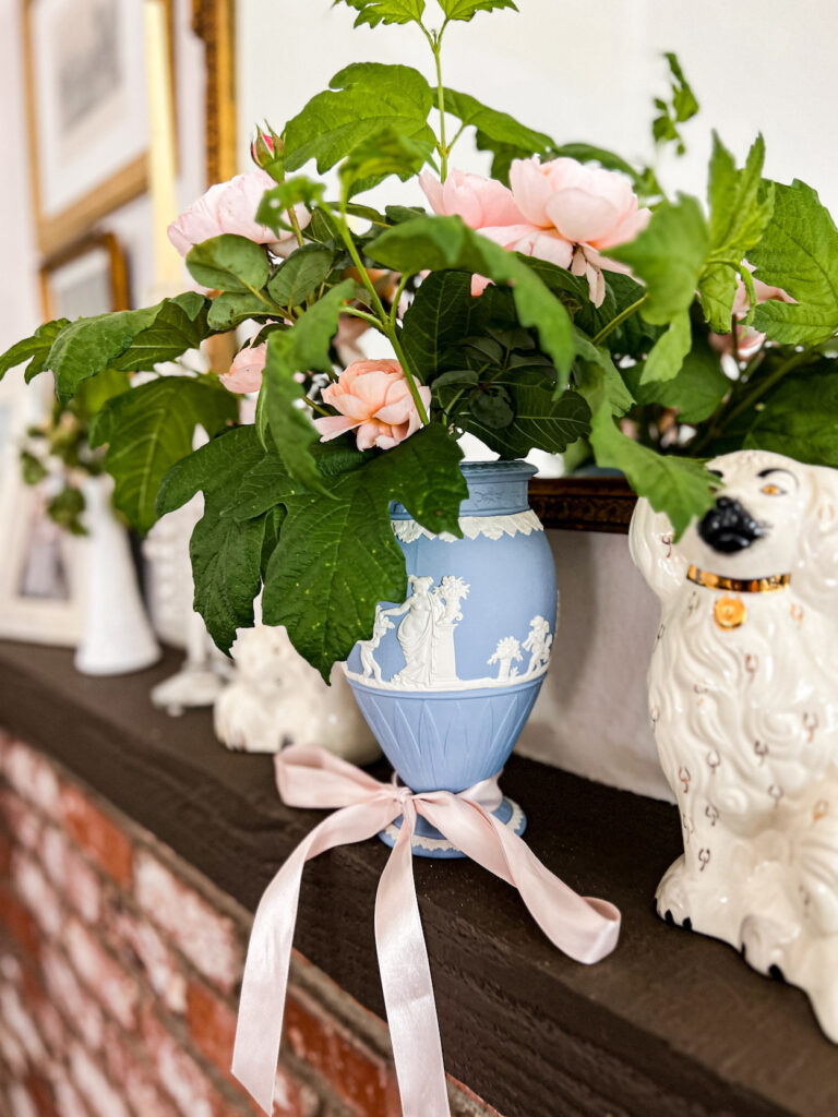 A side angle photo of a light blue and white Jasperware vase full of pink roses and greenery sits on a dark brown mantel between two white Staffordshire dogs (the dogs are slightly blurred and cropped out).