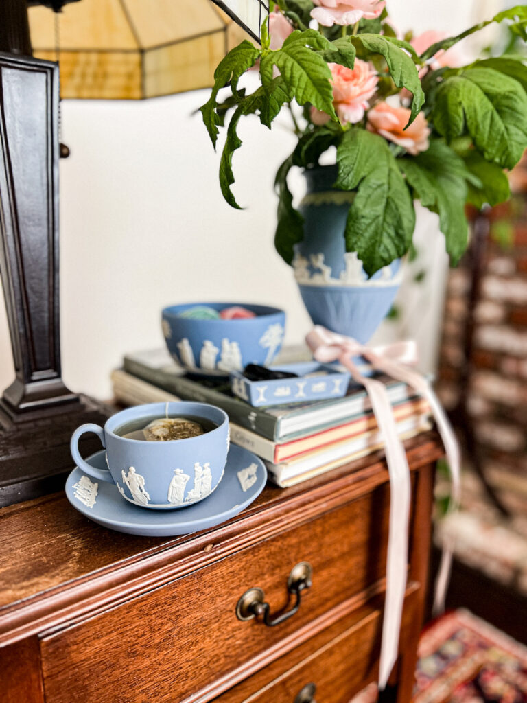 Four pieces of Jasperware--a teacup with a saucer, a bowl, a small lidless box, and a large vase--sit together on a side table. Each piece is the classic light blue (known officially as "lavender") with white accents. The teacup is in focus in the foregroud, while everything else is blurred in the background.