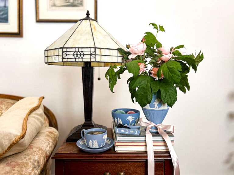 Four pieces of Jasperware--a teacup with a saucer, a bowl, a small lidless box, and a large vase--sit together on a side table. Each piece is the classic light blue (known officially as "lavender") with white accents.