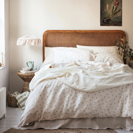 A wicker bed piled high with fluffy white linens with a soft floral print on the duvet cover.