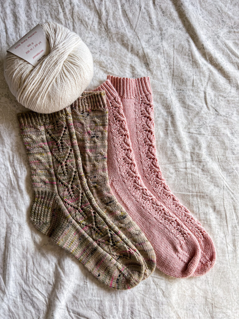 Two pairs of handknit socks, one green with pink speckles and one solid pink, are laid out on a bedsheet. A ball of white yarn fills the top left corner.
