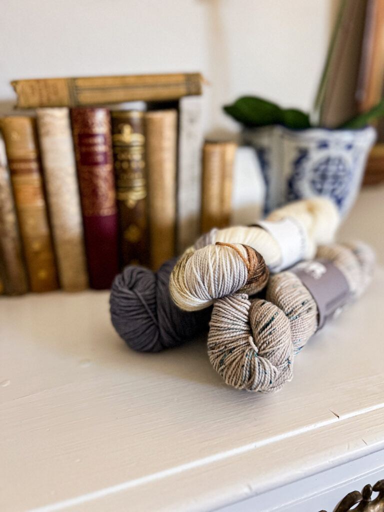 A pile of blue, gray, and white yarns are stacked on top of a white dresser. In the background are lots of antique books with gilded writing on their spines.