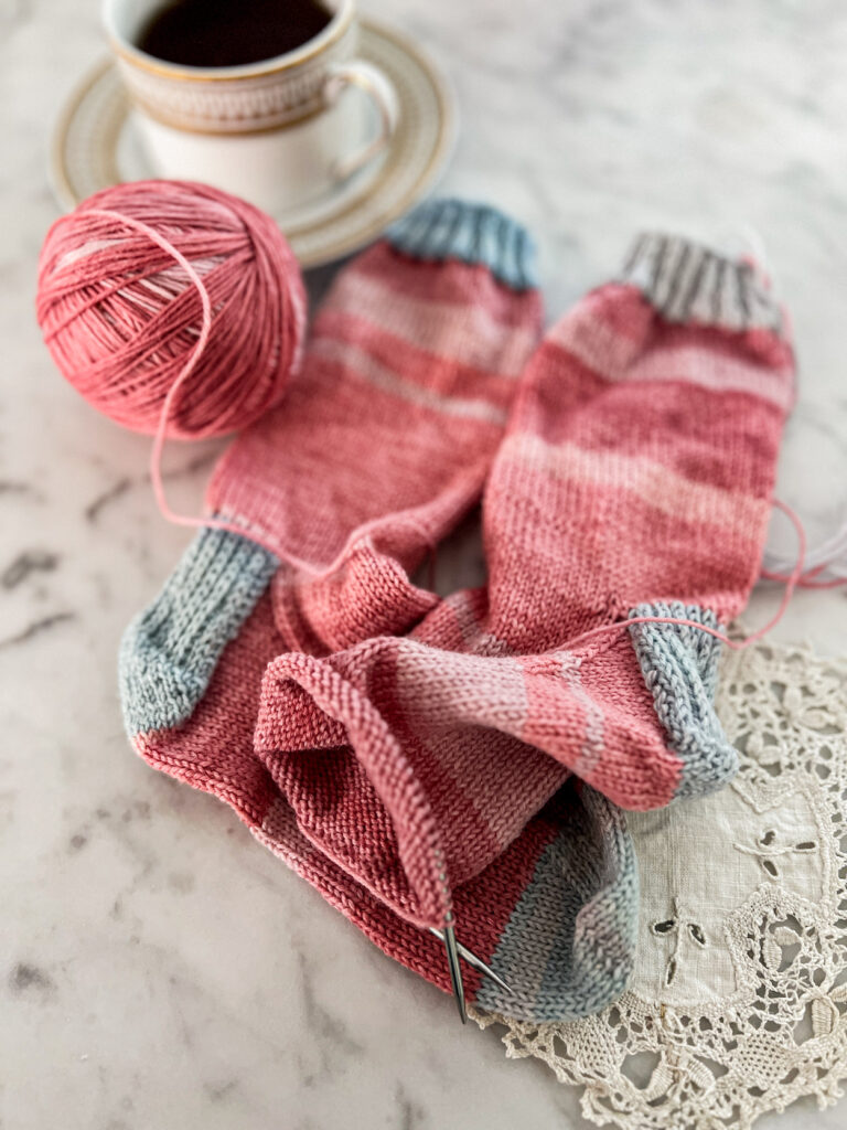 A low-angle photo focusing on the toes of a pair of pink handknit socks with contrasting blue cuffs, heels, and toes. The right sock is still on the needles and needs to be finished. Blurred in the background is a gold and white cup full of tea.